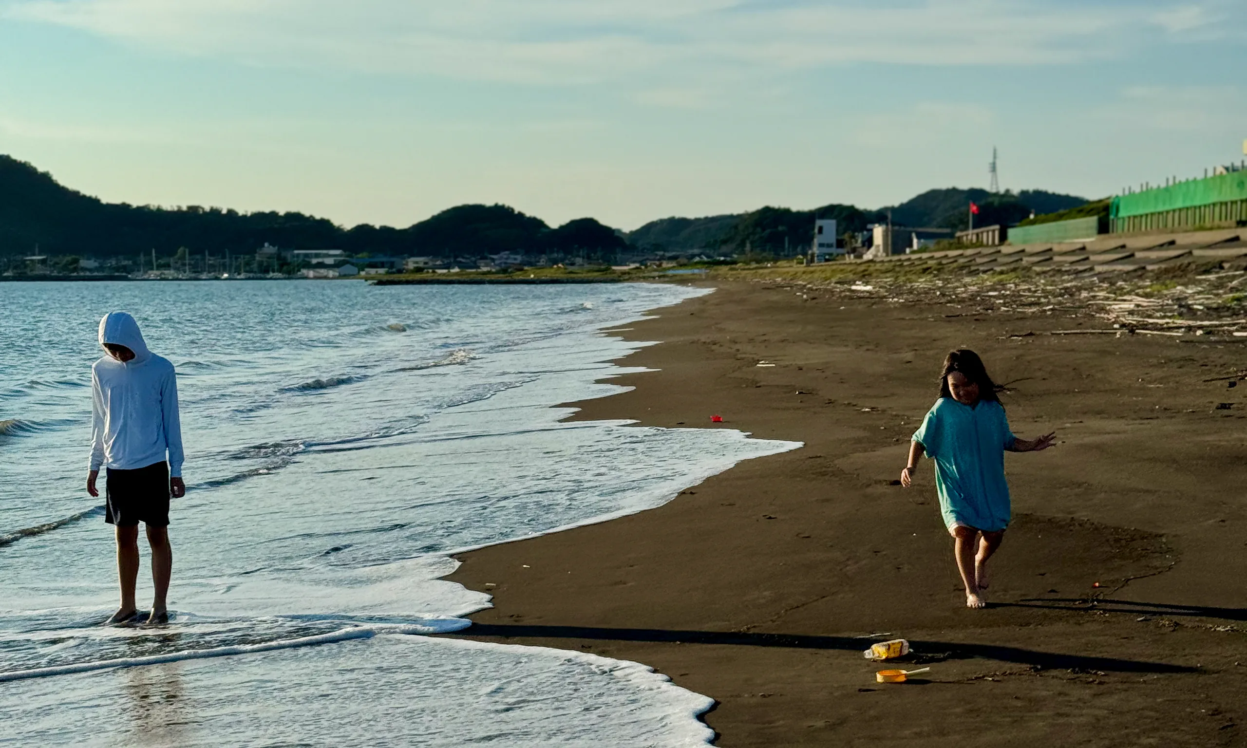 夕方の海辺。白いパーカーの子が波打ち際に立ち、水色の服の子が砂浜を走っている
